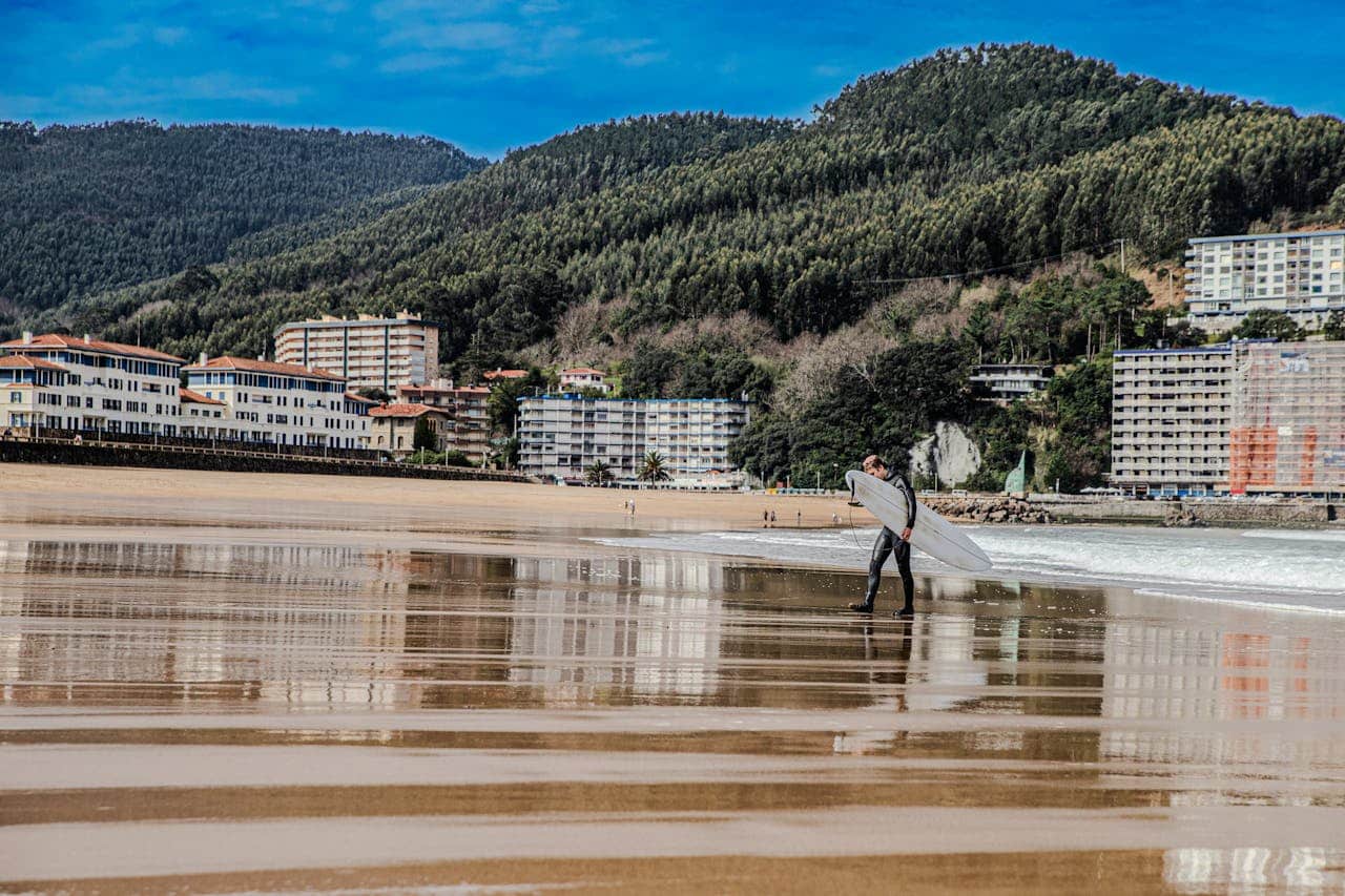 about-06 Surfer with board walking along Bakio Beach, surrounded by scenic buildings and hills.